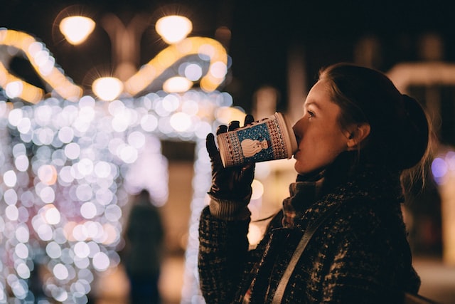 kvinde drikker kaffe foran en stor lys-dekoration som forestiller en gaveæske med sløjfe - kvinder passer på sit overskud i julen